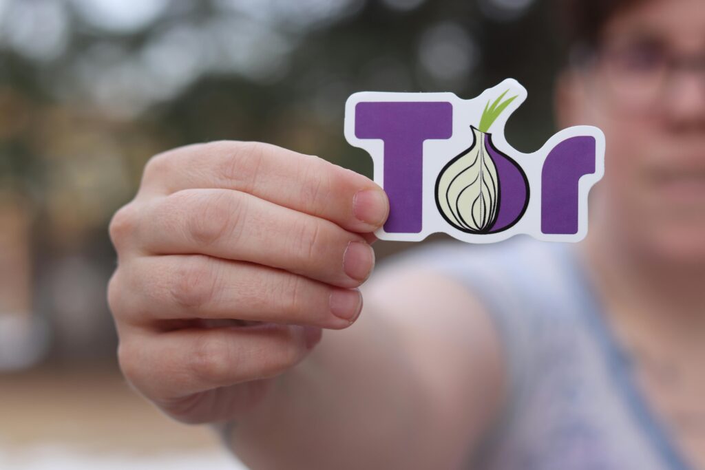 A close-up of a person holding a Tor browser logo sticker with a blurred outdoor background.